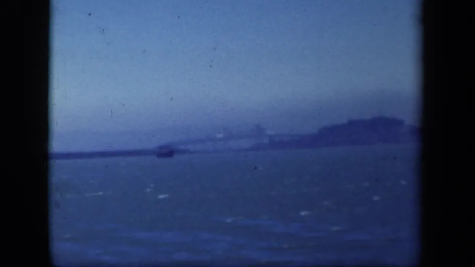 Distant San Francisco Bay home-movie view with choppy water, a small boat, an island form, and a bridge line in haze.