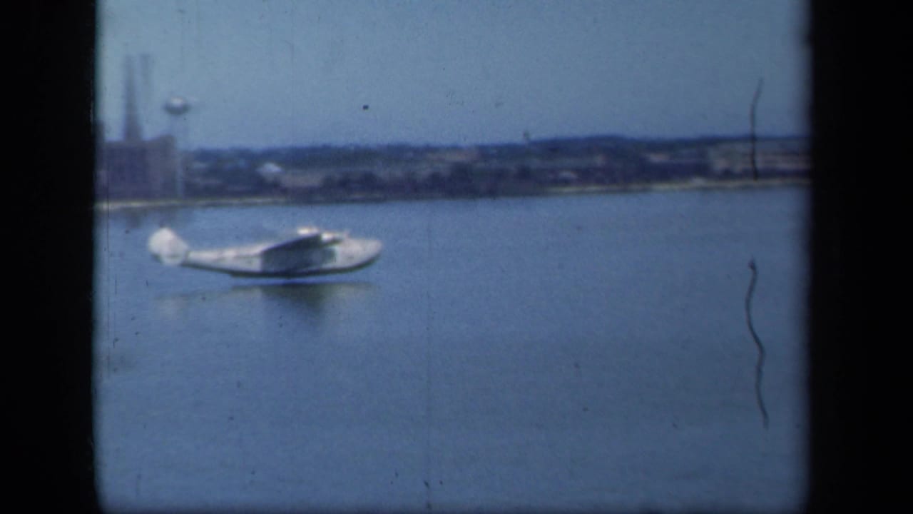 White seaplane glides across calm water with a low industrial shoreline in the background, filmed on 8mm in 1941.
