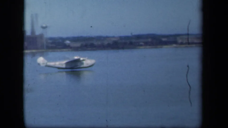 White seaplane glides across calm water with a low industrial shoreline in the background, filmed on 8mm in 1941.