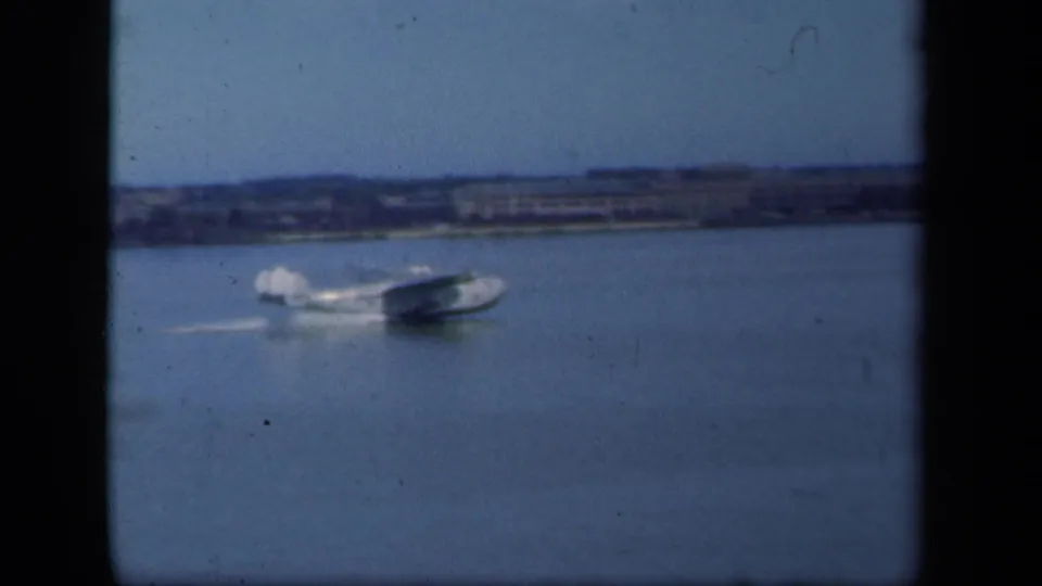 The seaplane accelerates across the water, leaving a white wake against a hazy 1941 waterfront backdrop.
