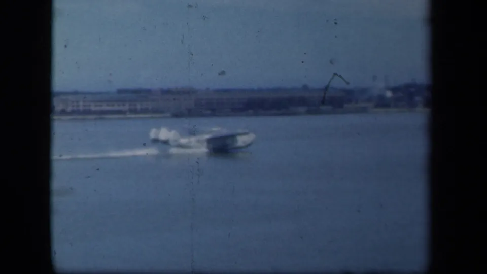 Seaplane continues across the water with a trailing wake, framed by dark 8mm gate edges and a distant New York-area shoreline.