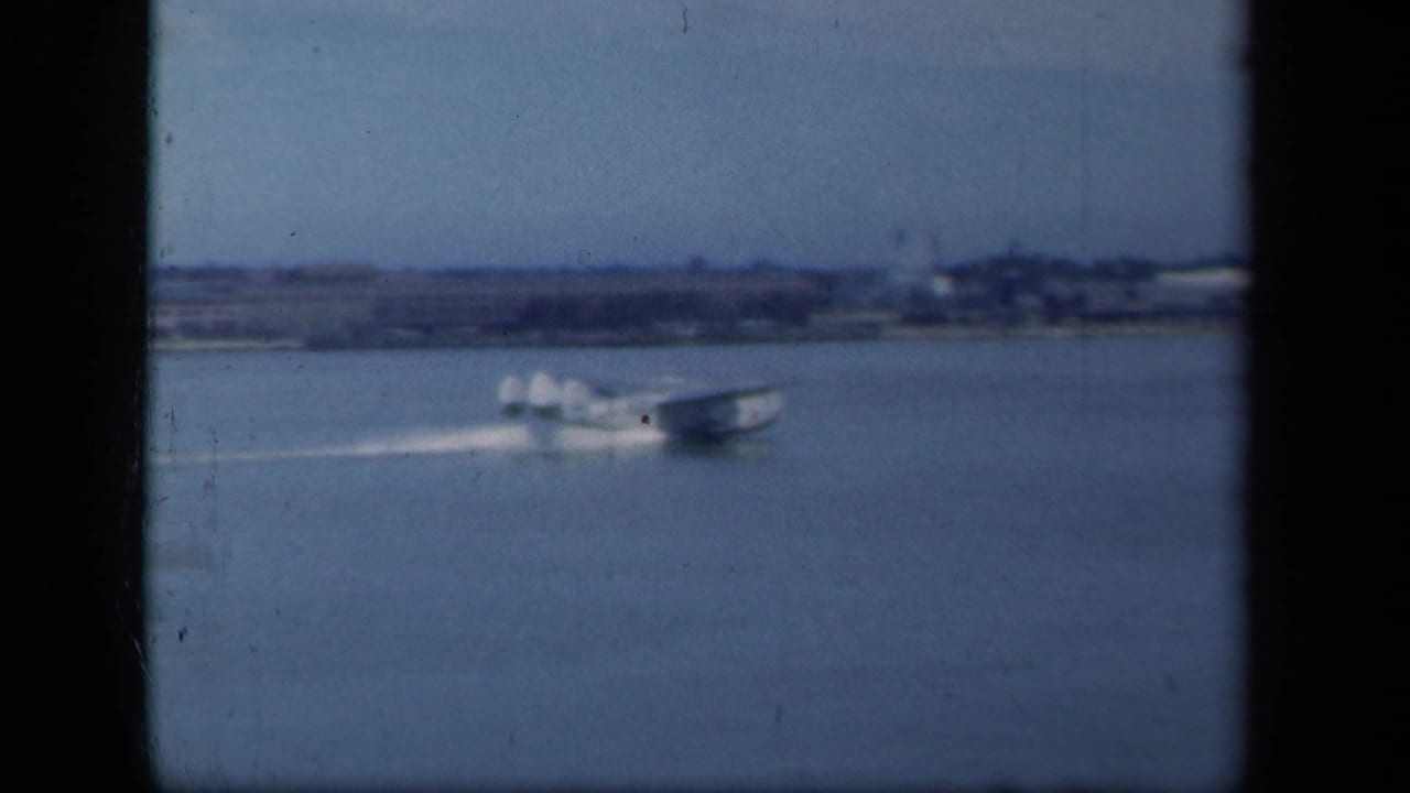 A 1941 New York home-movie frame shows a seaplane gliding across water, leaving a pale wake near a distant shoreline.