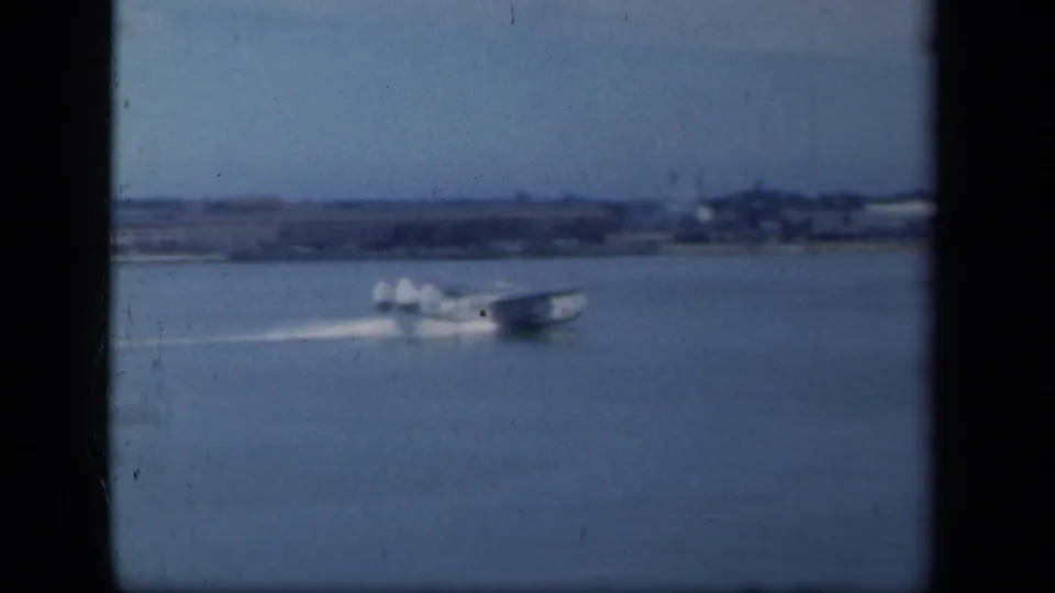 A 1941 New York home-movie frame shows a seaplane gliding across water, leaving a pale wake near a distant shoreline.