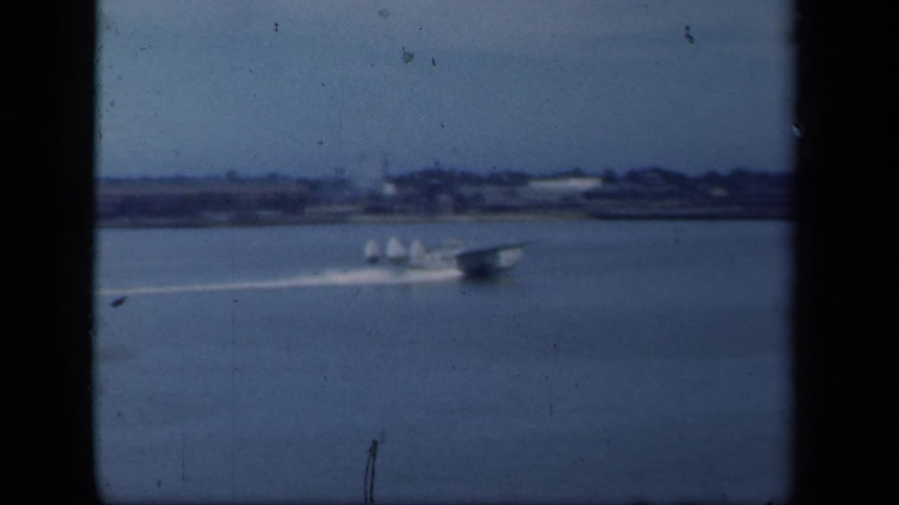 The seaplane continues across the New York waterfront, with a low shoreline visible through soft blue 8mm film color.