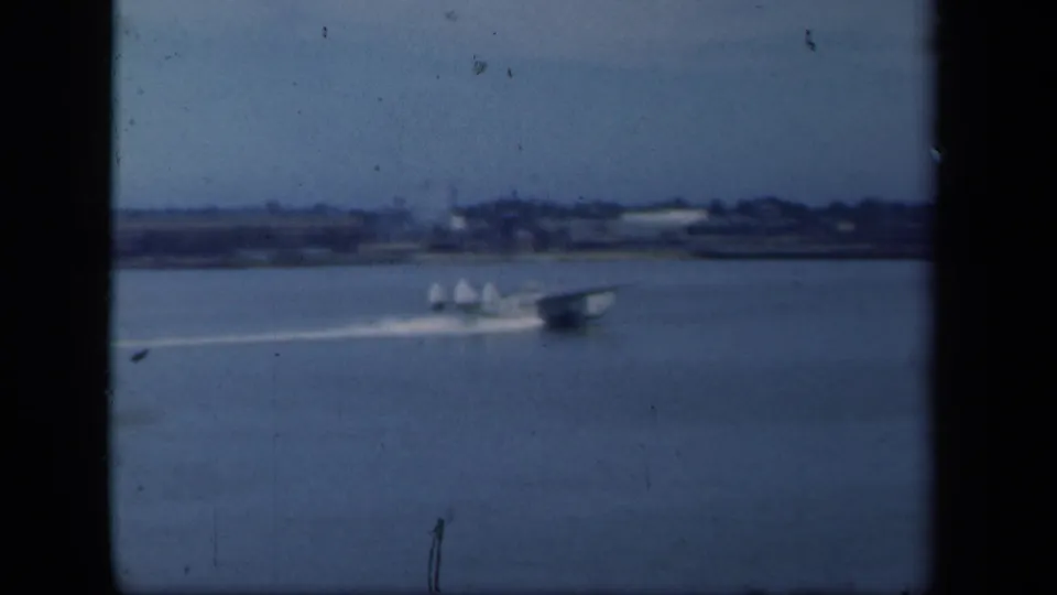 The seaplane continues across the New York waterfront, with a low shoreline visible through soft blue 8mm film color.