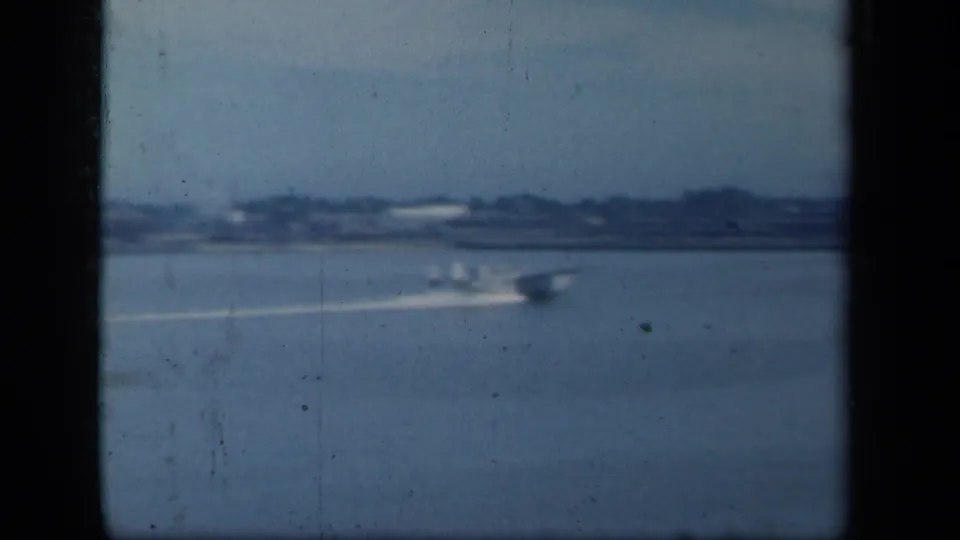 A soft-focus 8mm frame shows the seaplane and its white wake crossing water near a distant New York shoreline.