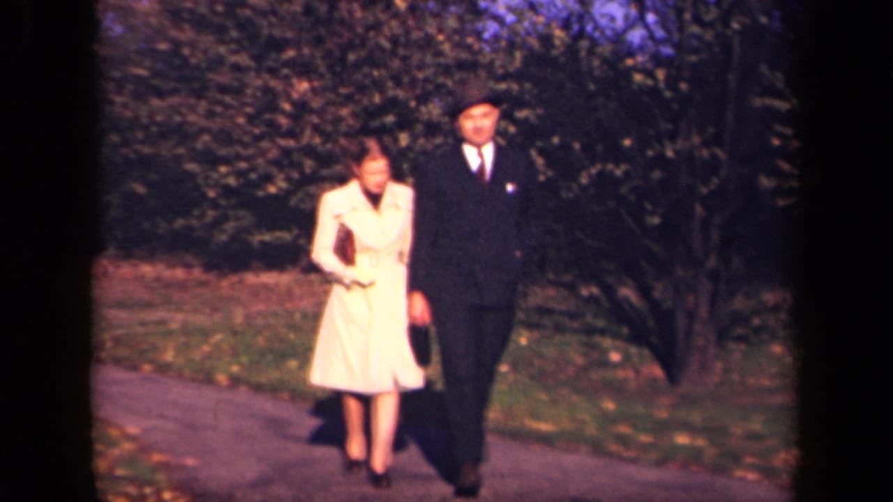 A woman in a light coat and a man in a dark suit walk closer on a park path.