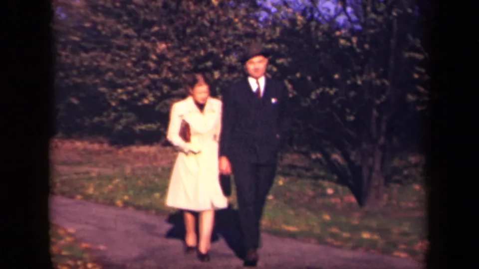 The couple continues toward the camera in a New York park, with the woman carrying a small purse and the man holding a hat.