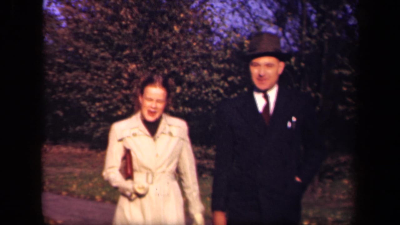 Woman in a light coat and man in a dark suit walk along a wooded path.