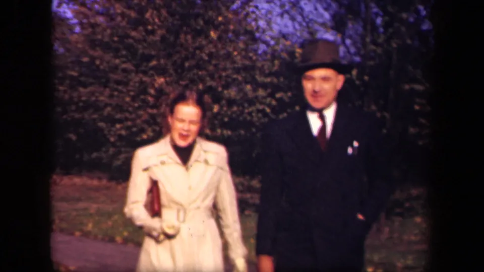 In 1941 New York home-movie footage, a woman in a light coat and a man in a dark suit and fedora walk toward the camera on a tree-lined path.