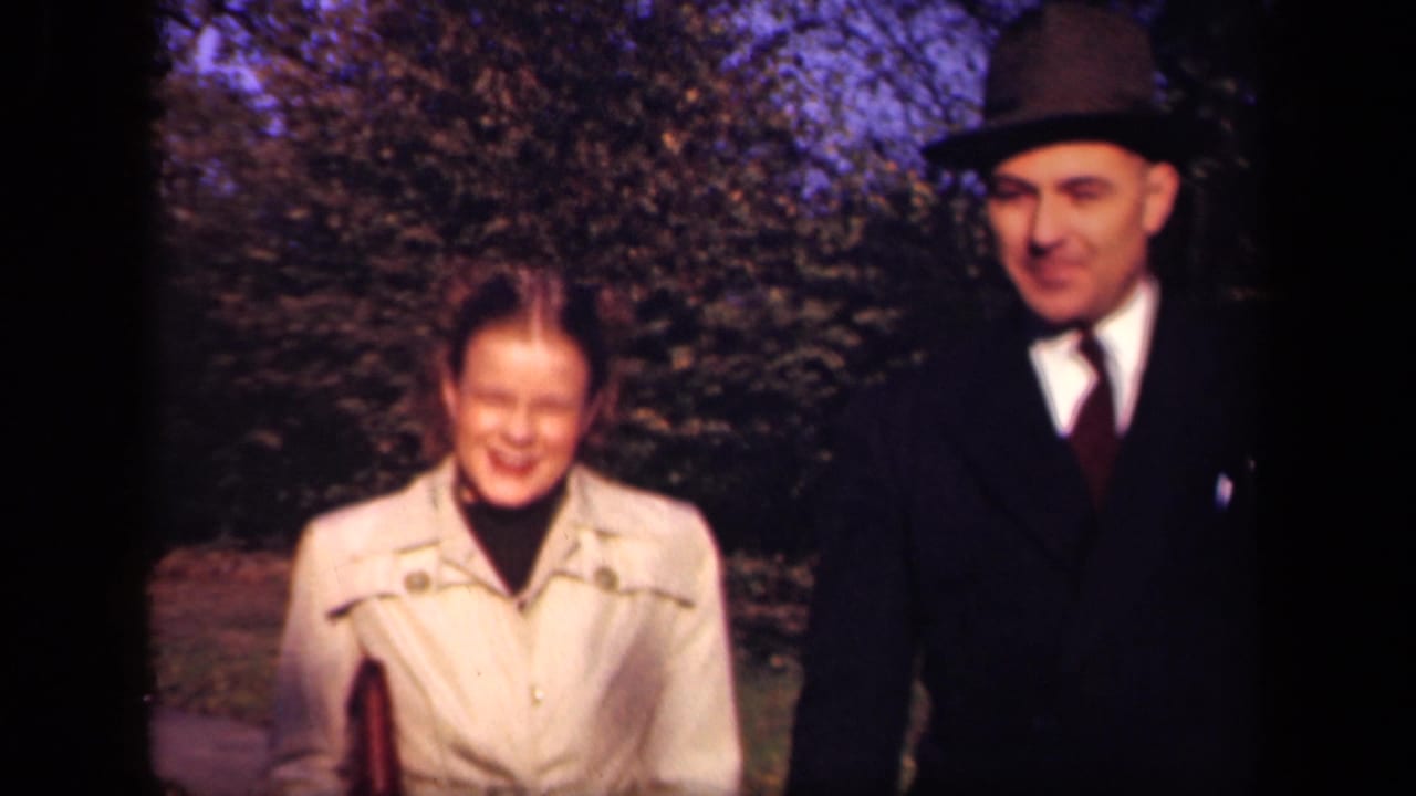 Closer 1941 New York frame of a woman in a light trench-style coat walking beside a man in a dark suit and fedora on an outdoor path.