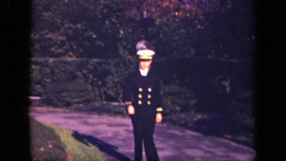 A young man in a formal navy-style uniform with gold buttons and peaked cap walks along a landscaped path in 1941 New York footage.