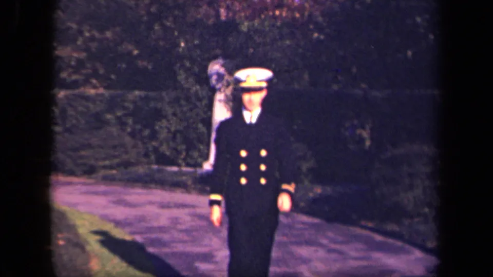 The navy-style officer continues toward the camera on a tree-lined path, with a second figure faintly visible behind him in the background.