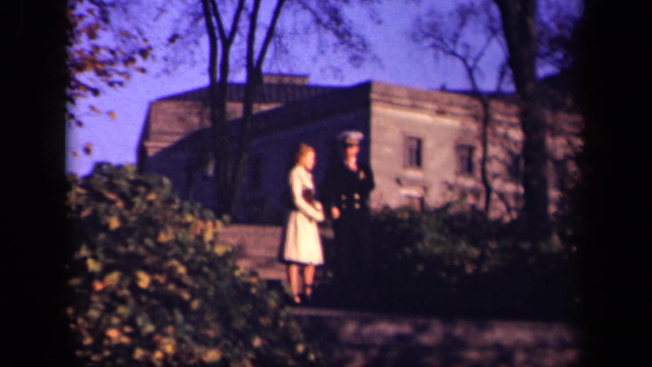 Uniformed man and woman in a pale dress stand on a path near a stone building.