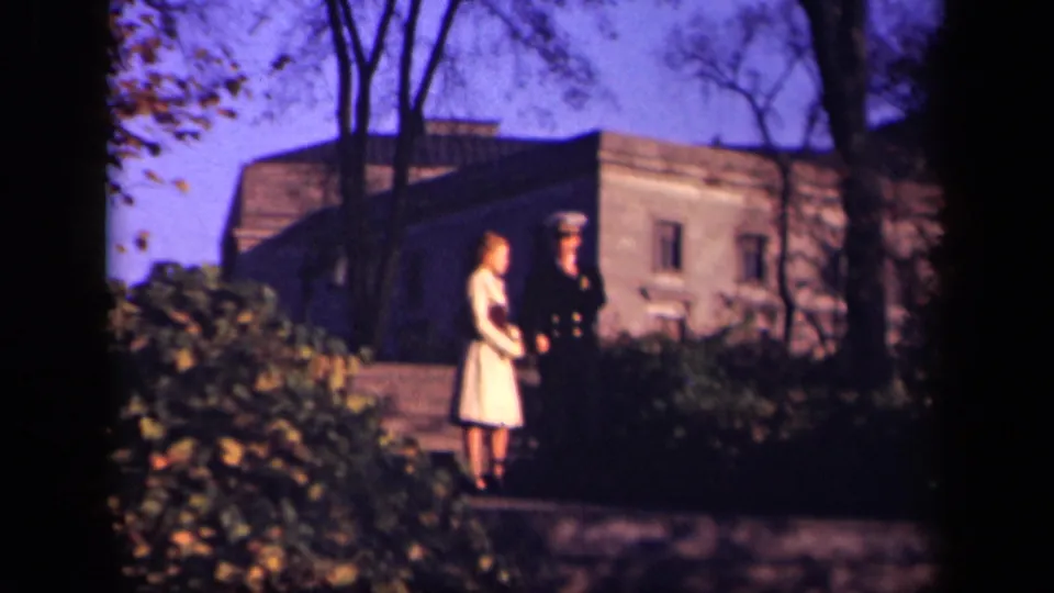 In 1941 New York home-movie footage, a uniformed man and a woman in a pale dress stand together outdoors near shrubs and a stone building.