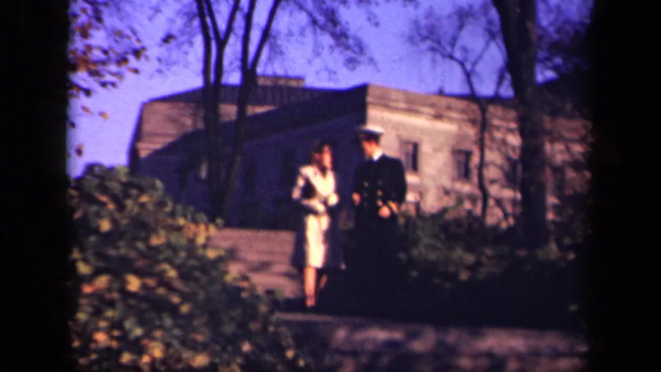 A uniformed man and woman in a pale dress approach the camera along a path, framed by trees and greenery in 1941 New York footage.