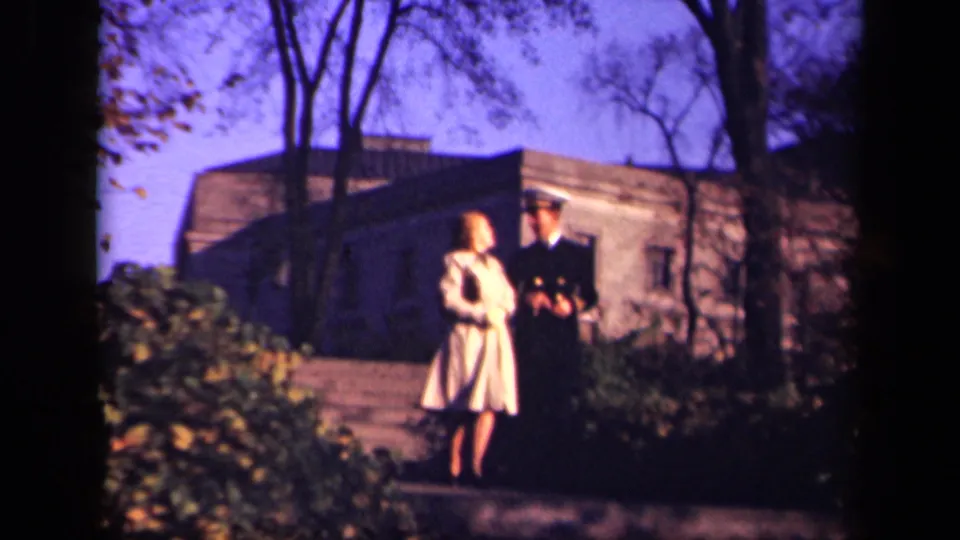 The woman and uniformed man stand near the camera on the path, with a low stone building and trees visible behind them in New York footage.