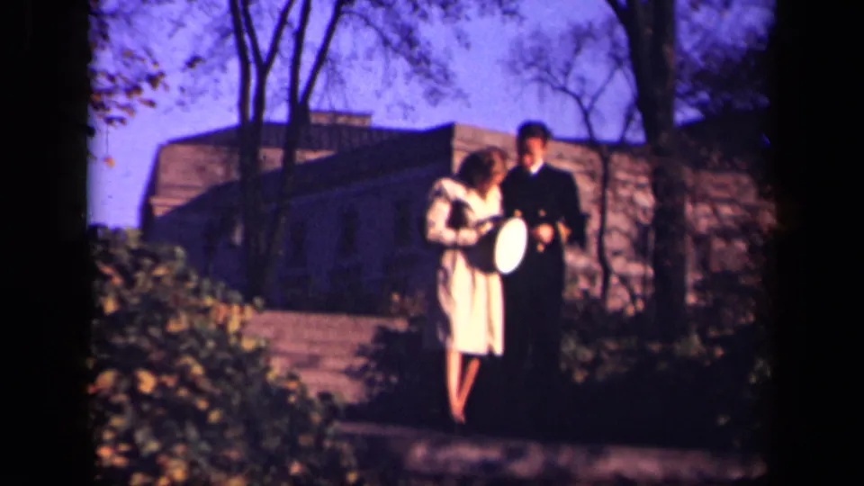 The young woman and uniformed man walk close together outdoors in the 1941 New York home-movie scene, with the pale hat visible as the stone building and trees remain behind them.