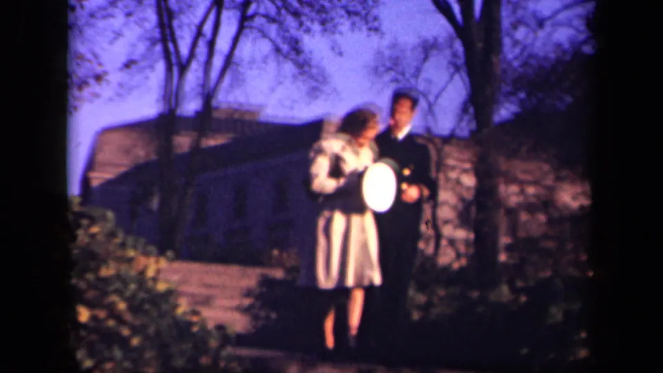 At the end of the 1941 New York clip, the woman looks down toward the light-colored hat while the uniformed man stands beside her against shrubs, trees, and the stone structure.