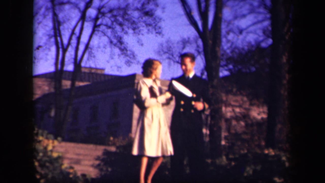 Woman in a light coat walks beside a man in a dark military uniform near trees and steps.