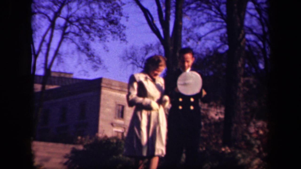 The uniformed man holds or adjusts his cap while the woman looks down beside him in the 1941 New York outdoor home-movie scene.