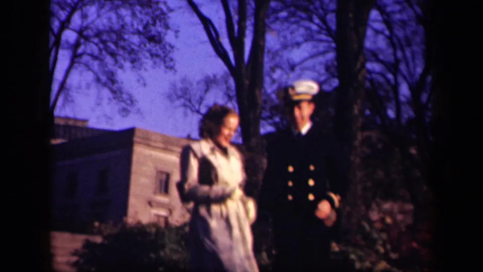 The couple continues walking outdoors, with the man now wearing a white cap and the woman in a light belted coat near shrubs and bare trees.