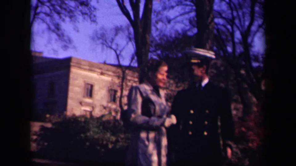The woman turns toward the uniformed man as they walk beside bare trees and a masonry building in the 1941 New York home movie.