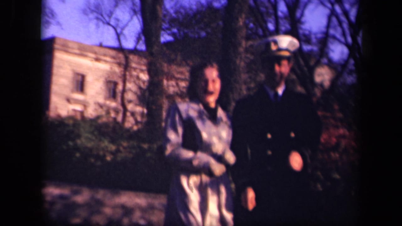 Woman in a light coat stands beside a man in a dark naval uniform outdoors.