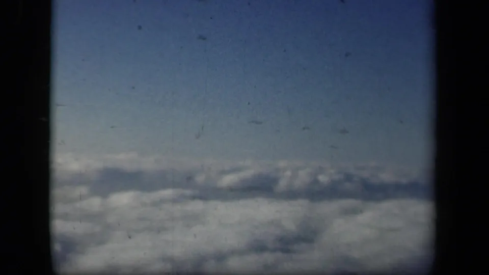 A broad layer of clouds appears below a blue sky, framed by the dark edges of an airplane window or cabin opening.