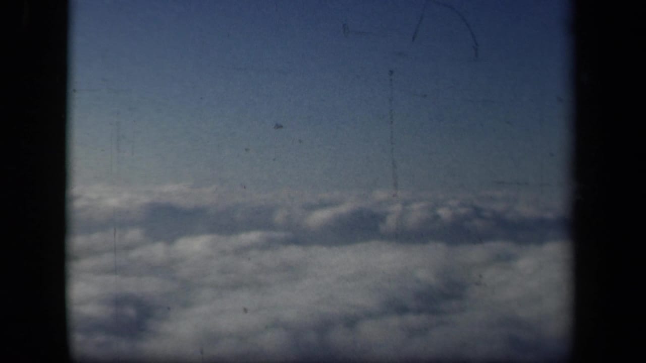 1941 8mm passenger view over clouds, framed by dark airplane window dividers with blue sky above.