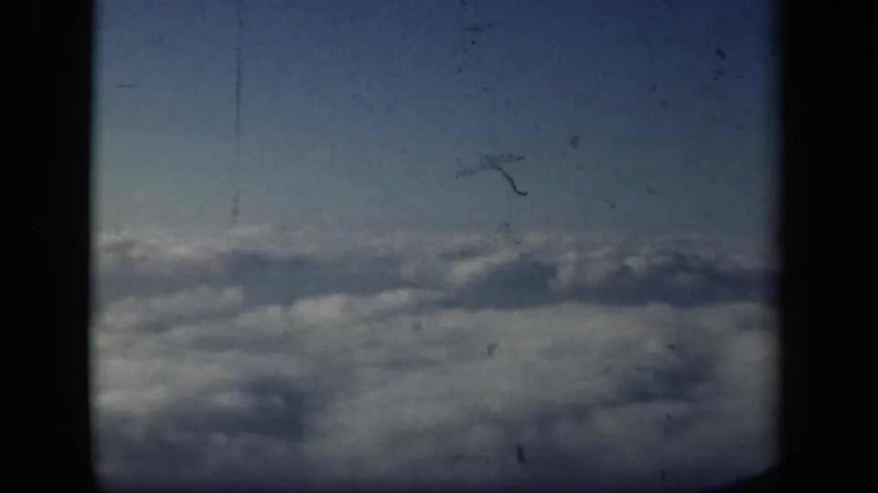 Home-movie airplane-window view over a layered cloud field, with 8mm grain and scratches visible across the frame.