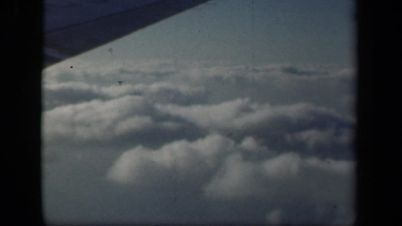 1941 8mm frame of soft cloud layers beneath a blue-gray sky, viewed past the wing edge from a passenger airplane window.