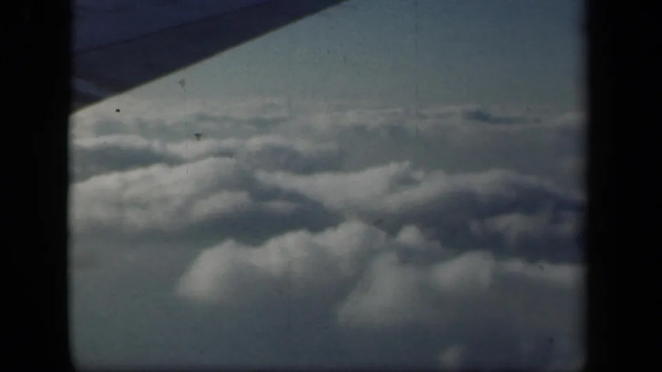 1941 8mm frame of soft cloud layers beneath a blue-gray sky, viewed past the wing edge from a passenger airplane window.