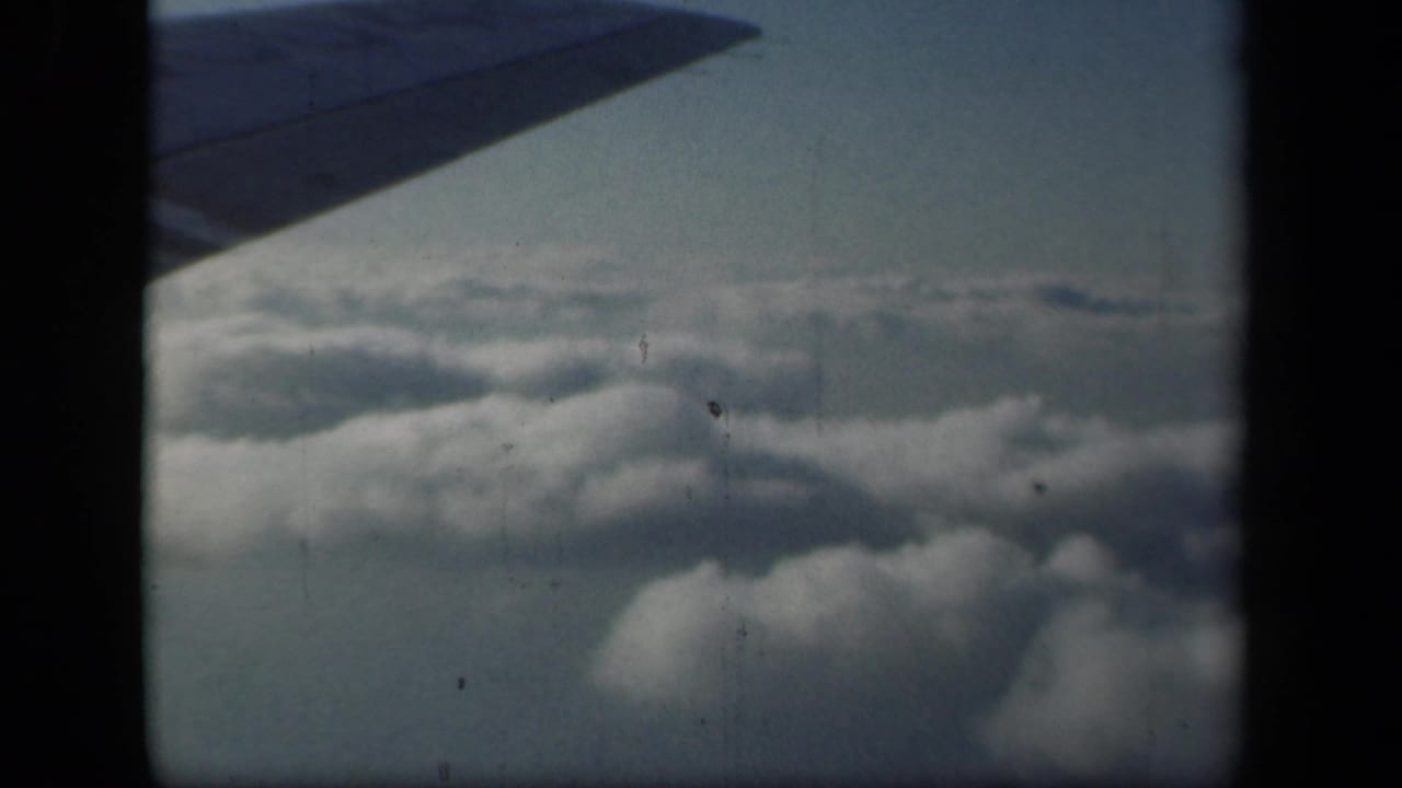 1941 8mm airplane-window view showing a dark wing silhouette over clouds, with soft color and visible film borders.
