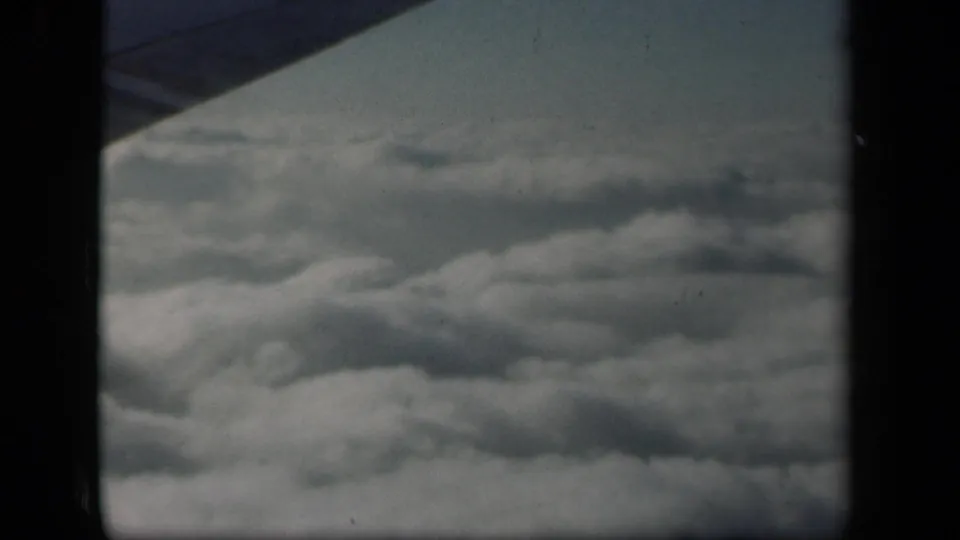 Soft 8mm passenger view above clouds, with a dark aircraft wing near the top edge and no ground visible.