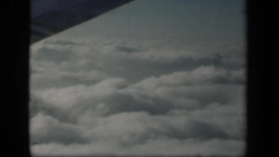 1941 home-movie frame looking out from an airplane over clouds, with the wing silhouetted against a pale sky.
