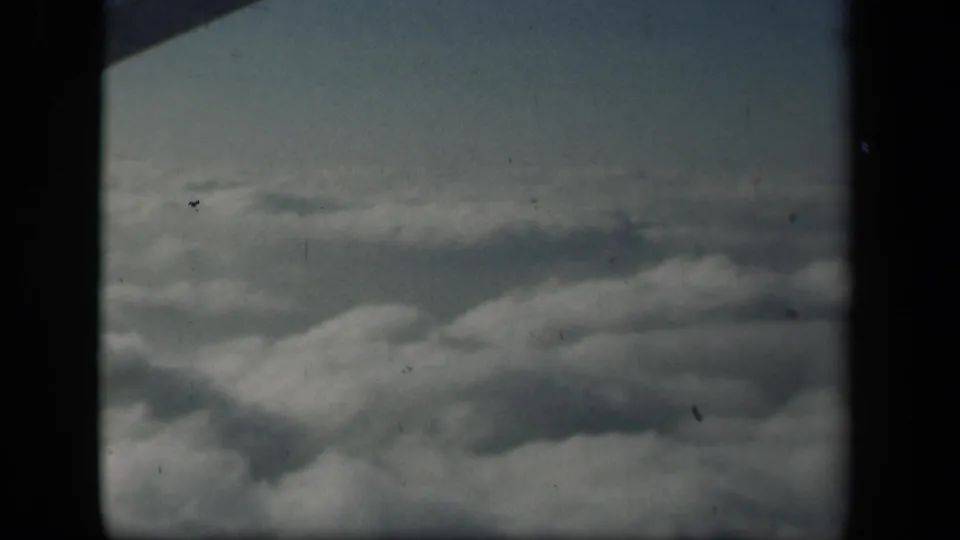 1941 8mm view from an airplane over puffy cloud tops, with visible grain, dust, and dark framing at the sides.