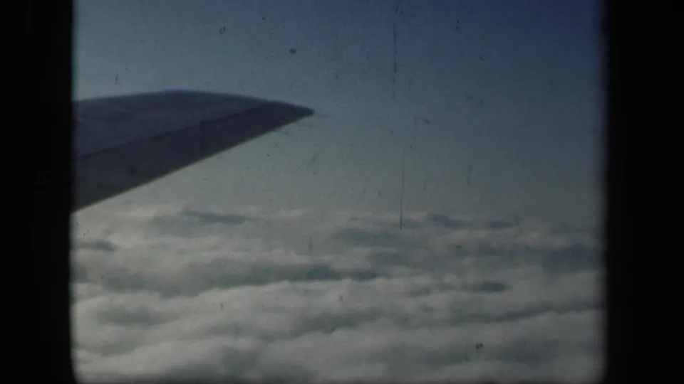 1941 home-movie frame from an airplane window, with wing silhouette, distant clouds, blue sky, and visible 8mm scan texture.