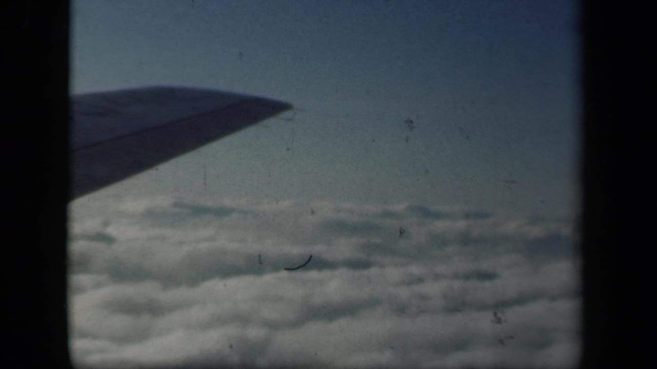 1941 8mm home-movie frame showing an airplane wing over a cloud deck, with blue sky and dark film borders.