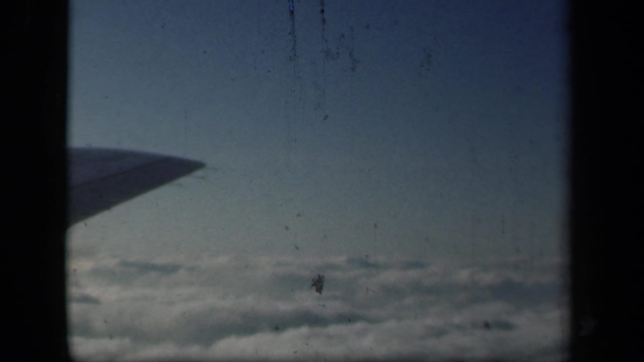 Airplane wing seen through a dark window frame above clouds and blue sky.