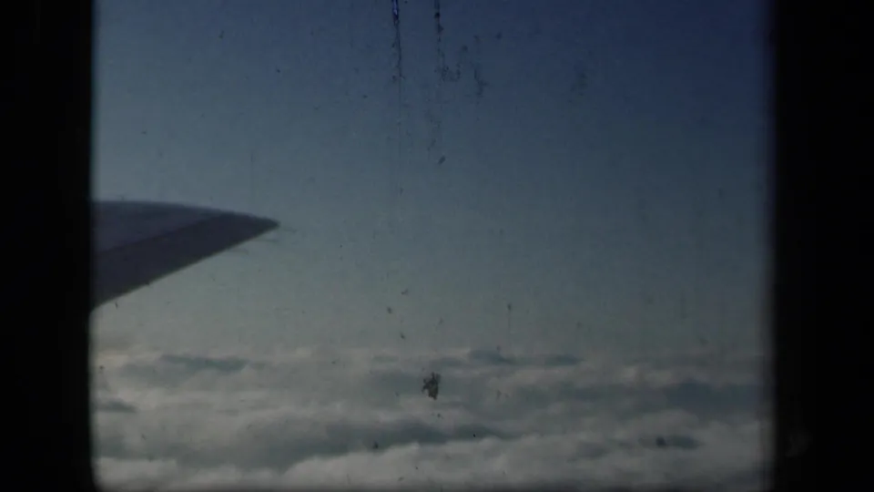 1941 8mm passenger view from an airplane window, with the wing crossing blue sky above a cloud layer.