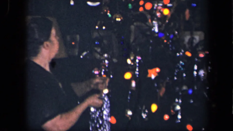 The home-movie frame shows the woman turned toward the Christmas tree, holding tinsel among glowing colored bulbs and ornaments.