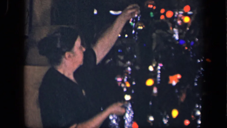 A woman reaches up to place silver tinsel high on a Christmas tree, with colored lights visible in the dim 1942 home interior.