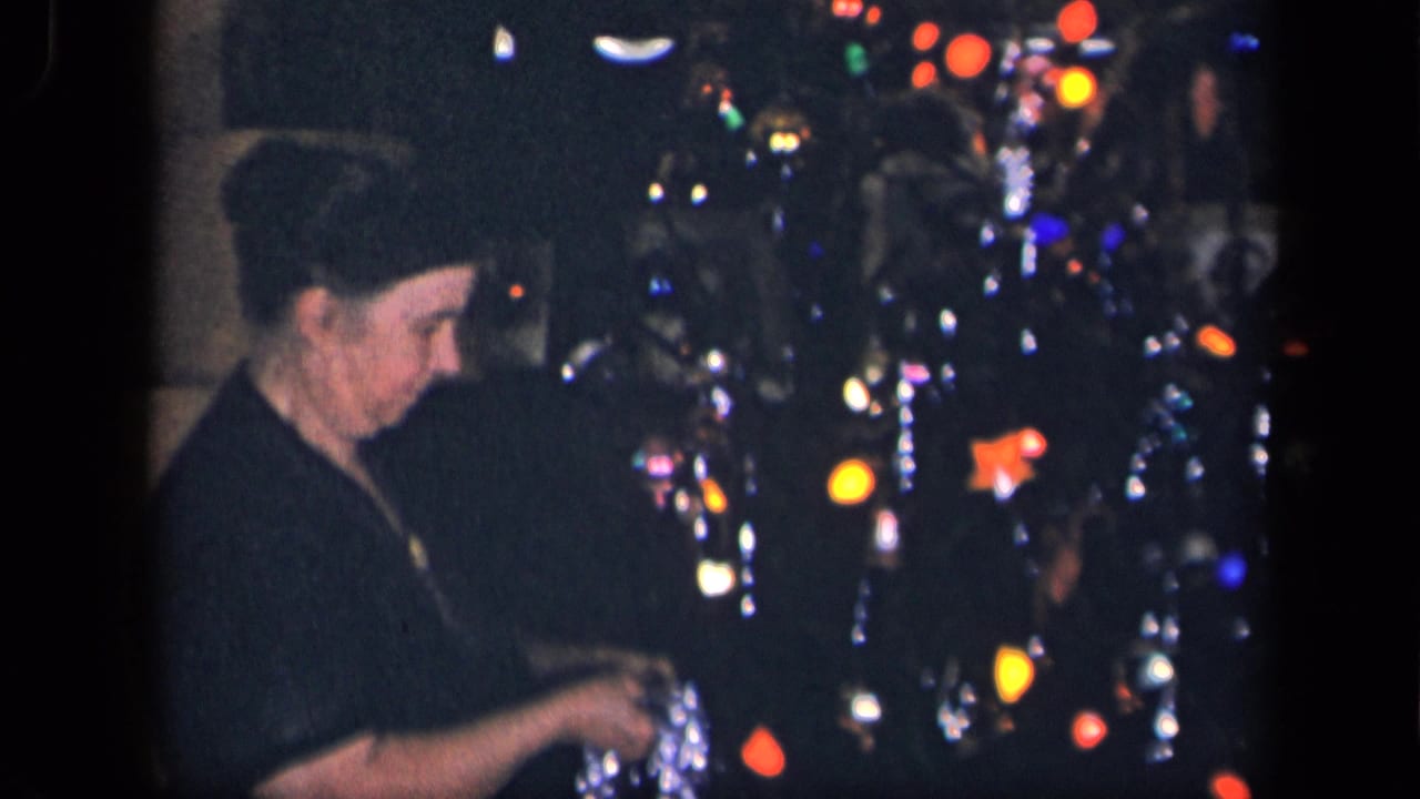 An older woman in a dark dress stands near a decorated Christmas tree with colored bulbs and hanging tinsel in a private interior.
