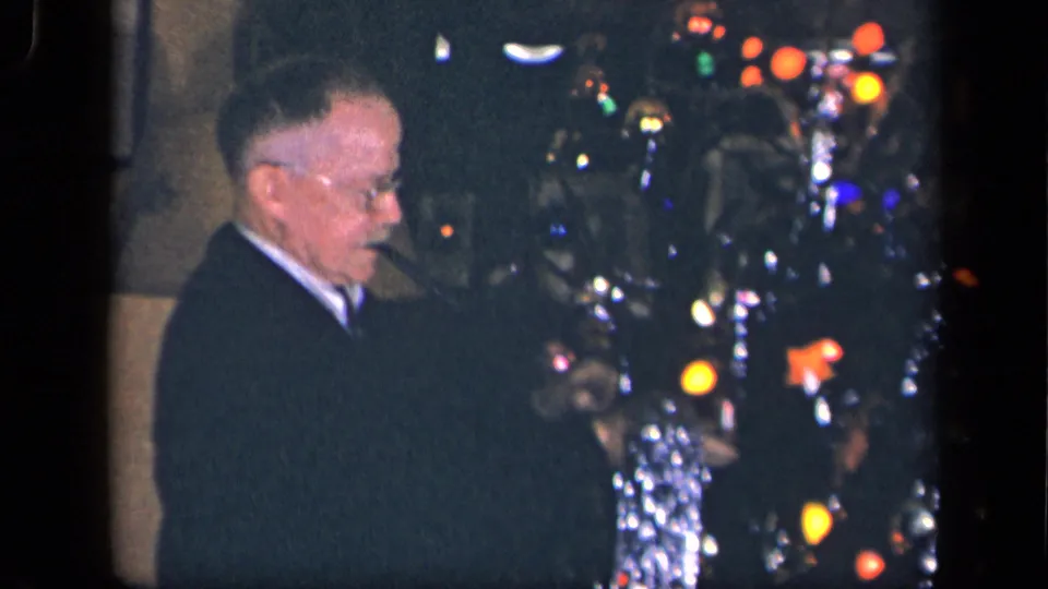 An older man wearing glasses and a dark suit stands by the Christmas tree, holding a pipe in supported 1942 Sebring, Ohio footage.