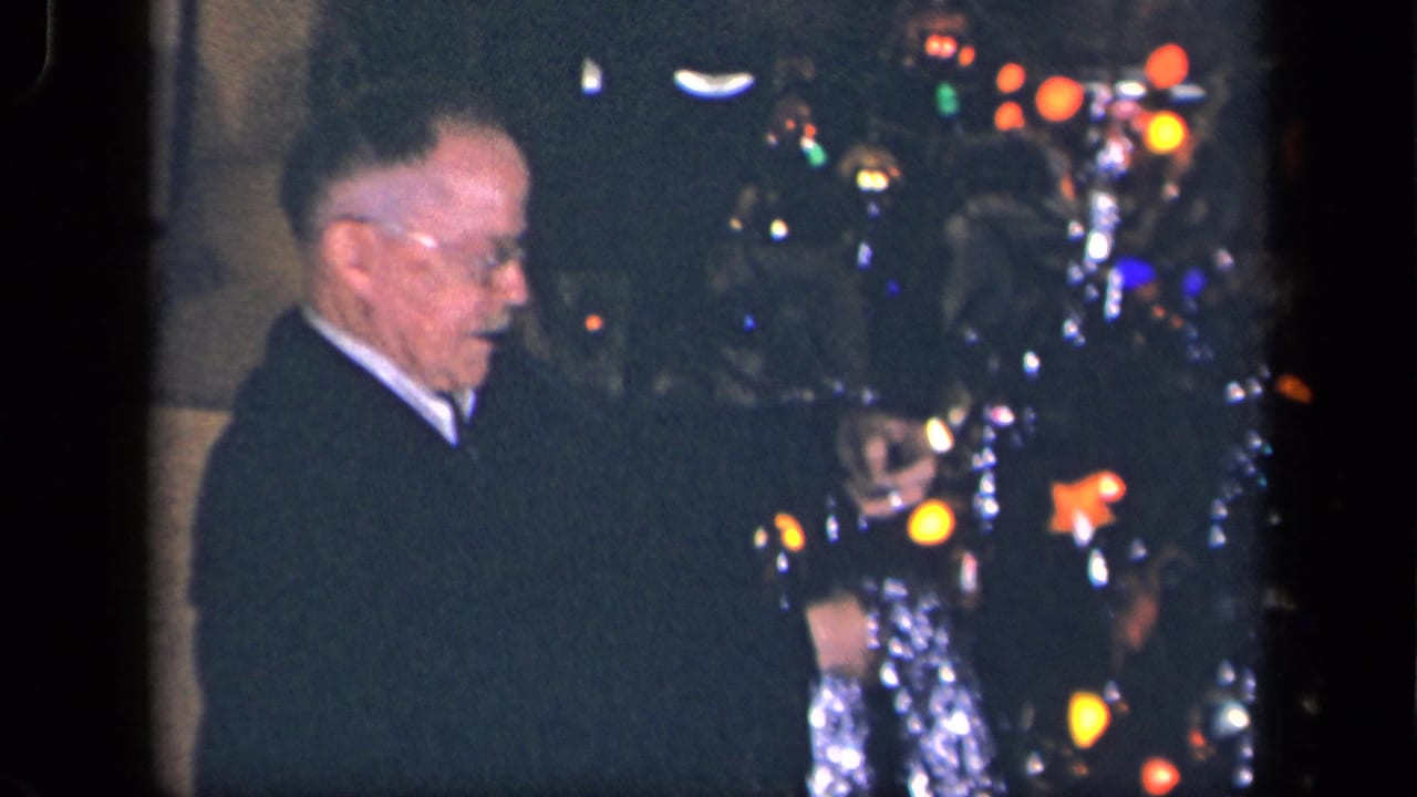 In 1942 Sebring, Ohio home-movie footage, an elderly man in a dark suit stands near a lit Christmas tree with tinsel and ornaments.