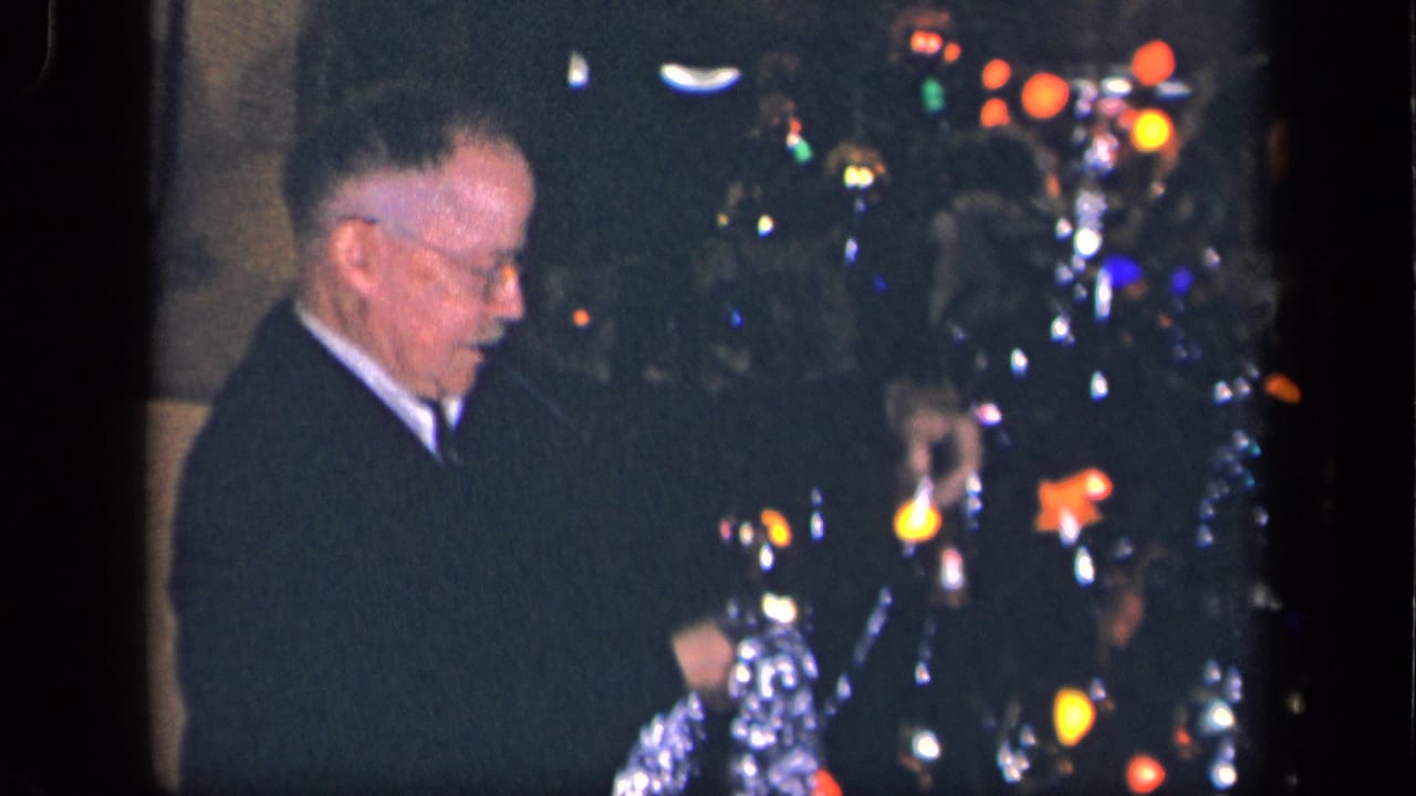 The elderly man faces the Christmas tree as colored bulbs and silver tinsel glow in the dim 1942 Sebring, Ohio interior.