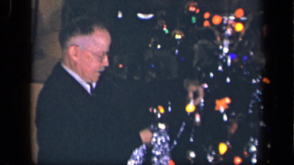 The elderly man faces the Christmas tree as colored bulbs and silver tinsel glow in the dim 1942 Sebring, Ohio interior.