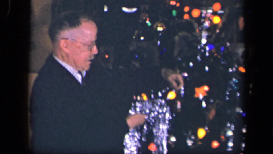 An elderly man in glasses holds or arranges silver tinsel beside a lighted Christmas tree in the Sebring, Ohio home-movie scene.