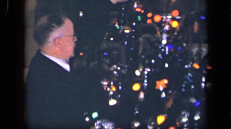 In a 1942 Sebring, Ohio home-movie frame, an older man in glasses faces a decorated Christmas tree with colored lights and tinsel.